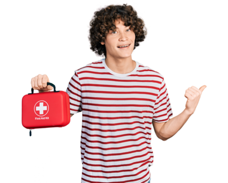 Caucasian teenager holding first aid kit pointing thumb up to the side smiling happy with open mouth