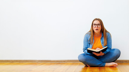 Surprised woman with glasses sits on the floor against white wall holding book