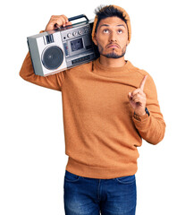 Handsome latin american young man holding boombox, listening to music pointing up looking sad and...
