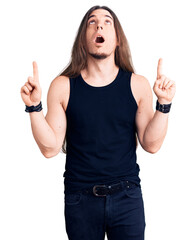 Young adult man with long hair wearing goth style with black clothes amazed and surprised looking...