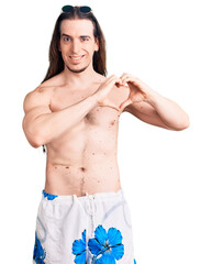 Young adult man with long hair wearing swimwear shirtless smiling in love showing heart symbol and shape with hands. romantic concept.