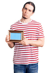 Young adult man with long hair holding empty frame thinking attitude and sober expression looking self confident