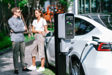 Young couple travel with EV electric car charging in green sustainable city outdoor garden in summer shows urban sustainability lifestyle by green clean rechargeable energy of electric vehicle innards - obrazy, fototapety, plakaty