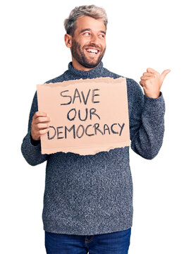 Young blond man holding save our democracy cardboard banner pointing thumb up to the side smiling happy with open mouth