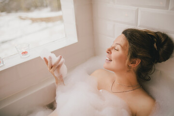 Young woman taking bath with foam near big window. - obrazy, fototapety, plakaty