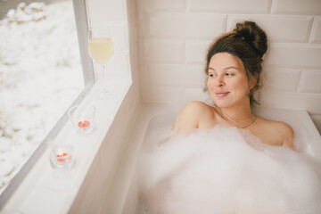 Young woman taking bath with foam near big window.
