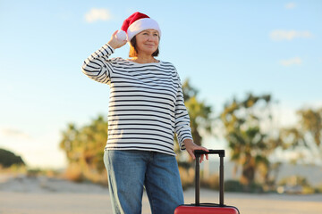 Cheerful mature woman 50s in Christmas Santa Claus hat, striped shirt holds red suitcase on beach...
