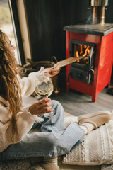 Young woman sitting by the fireplace in white sweater, drinking wine in cozy log cabin.