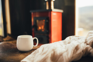 White cup with tea with burning fireplace on the background in cozy log cabin.
