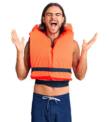 Young handsome man wearing nautical lifejacket celebrating mad and crazy for success with arms...