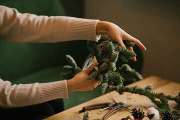 Woman making Christmas wreath using natural pine branches, sitting near wooden table.