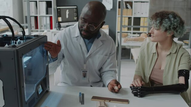 Zoom out shot of Black doctor describing operating principles of 3d printing machine while it creating new prosthetic for girl with disabilityZoom out shot of Black doctor describing operating princip