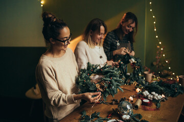 Happy women making Christmas wreath using natural pine branches and festive decorations.