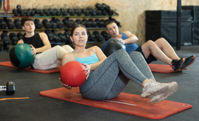 Young dedicated woman doing exercises with medicine ball near other people in gym