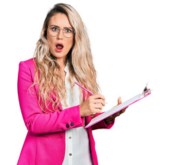 Young blonde woman writing on clipboard in shock face, looking skeptical and sarcastic, surprised...