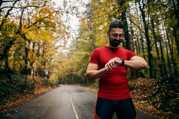 Fototapeta premium An athlete is standing on a rainy weather in nature and looking at achievement on a smart watch..