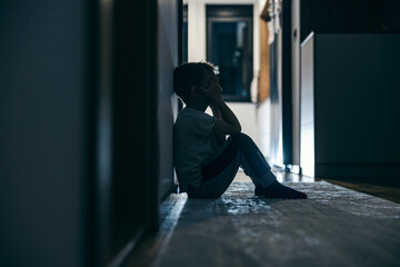 Side view of a crying boy sitting in a dark at home.