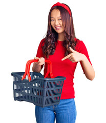 Young beautiful chinese girl holding supermarket shopping basket smiling happy pointing with hand and finger