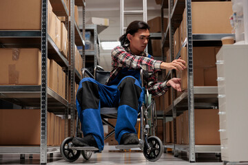 Warehouse asian package handler in wheelchair searching cardboard box on shelf. Shipment manager...