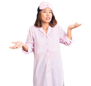 Young beautiful chinese girl wearing sleep mask and pajama clueless and confused expression with arms and hands raised. doubt concept.