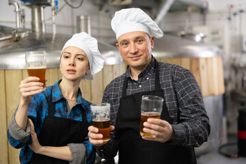 Interested concentrated male and female brewers wearing black aprons holding glasses of beer in hands, examining quality of beverage in craft brewery