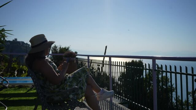 Silhouette of female tourist in hat and sunglasses using smartphone in Amalfi coast, taking photo, shooting video sitting chair on background of beautiful seascape, on summer sunny day, slow motion.