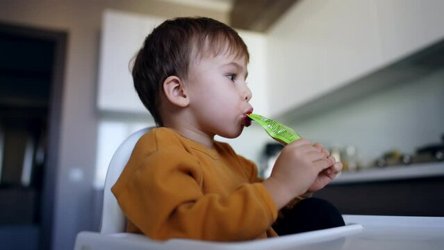 Little boy having a snack from the doy pack. Kid eating puree fruit at home. Low angle view.