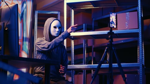 Hacker wearing anonymous mask doing ransom video after gathering information from users computers using spyware software. Cybercriminal blackmailing victim in graffiti painted hideout