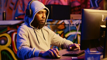 Hacker stealing credit card numbers and manipulating banking systems for his own personal gain while listening to music through headphones. Evil developer programming in graffiti painted basement