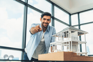 Portrait of smart engineer in casual outfit smiling at camera while holding pencil. Businessman pointing at camera and standing near house model, project plan with sky scraper behind. Tracery.