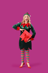 Happy young woman dressed as elf with Christmas gift on purple background