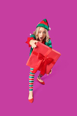 Emotional young woman dressed as elf with Christmas gift on purple background