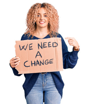 Young blonde woman with curly hair holding we need a change banner smiling happy pointing with hand and finger