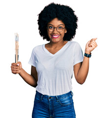 Young african american woman holding paintbrushes screaming proud, celebrating victory and success...