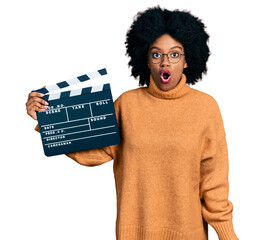 Young african american woman holding video film clapboard scared and amazed with open mouth for...