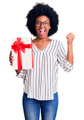 Young african american woman holding gift screaming proud, celebrating victory and success very excited with raised arms