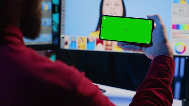 Photo editor using green screen mobile phone to watch tutorial, learning to color correct photographs in creative studio. Photographer watching guiding video on chroma key smartphone