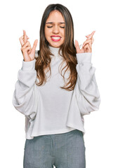 Young brunette woman wearing casual turtleneck sweater gesturing finger crossed smiling with hope and eyes closed. luck and superstitious concept.