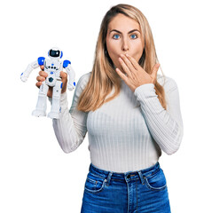 Young blonde woman holding robot toy covering mouth with hand, shocked and afraid for mistake....