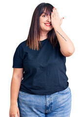 Young plus size woman wearing casual clothes covering one eye with hand, confident smile on face and surprise emotion.