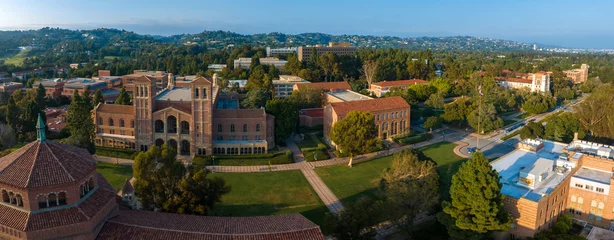 Fototapeten Aerial view of UCLA campus with Gothic tower, red-brick buildings, green lawns, and pathways amidst hilly, tree-covered landscape in soft, golden light. © ingusk