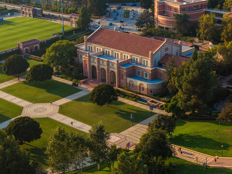 Aerial view of a grand university campus with red-bricked buildings, green lawns, and classical architecture, featuring a sports field, pond, and serene outdoor seating areas.