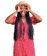 Young african american woman wearing summer hat doing ok gesture like binoculars sticking tongue out, eyes looking through fingers. crazy expression.