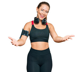 Beautiful blonde woman wearing gym clothes and using headphones smiling cheerful offering hands giving assistance and acceptance.
