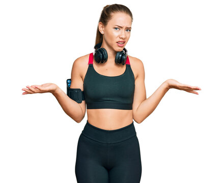 Beautiful blonde woman wearing gym clothes and using headphones clueless and confused with open arms, no idea concept.