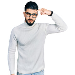 Young arab man with beard wearing elegant turtleneck sweater and glasses pointing unhappy to pimple...