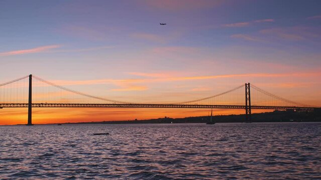 View of 25 de Abril Bridge famous tourist landmark of Lisbon connecting Lisboa and Almada on Setubal Peninsula over Tagus river with yacht silhouette on sunset and flying plane. Lisbon, Portugal