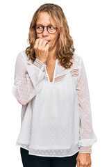 Middle age blonde woman wearing casual white shirt and glasses looking stressed and nervous with hands on mouth biting nails. anxiety problem.