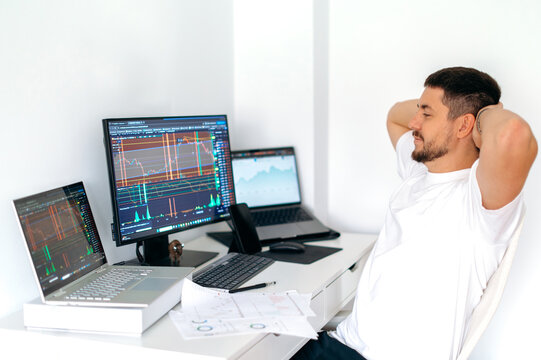 Stock trading, cryptocurrency concept. Male trader sitting at desk at home office monitoring stock market looking at monitors analyzing price flow, risks and prospects