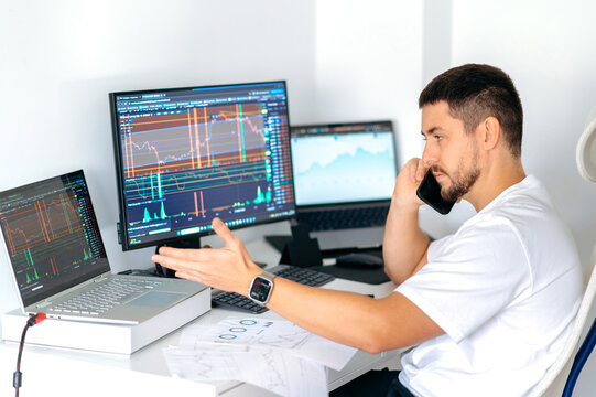 Focused male trader sits at desk at home office monitoring stock market looking at monitors analyzing price flow, risks and prospects while talking on the phone. Stock trading, cryptocurrency concept